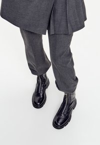 Persoană îmbrăcată în pantaloni gri închis și cizme de piele negre până la gleznă cu fermoar frontal, pe un fundal alb.
