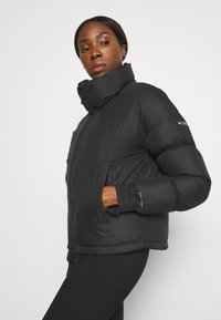 Svart puffjacka med hög krage, dragkedjestängning och elastiska ärmslut. Har två sidofickor och en slät, snygg yta.