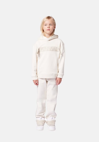 Felpa bianca con una fascia testurizzata e decorata sul petto, abbinata a pantaloni tuta coordinati e sneakers bianche con dettagli in maglia.