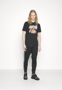 Fekete grafikai póló, melyen a "JORDAN" felirat és egy kosárlabdázó nyomat található, fekete sportos melegítőnadrággal, kék oldalsávokkal és fekete tornacipővel párosítva.