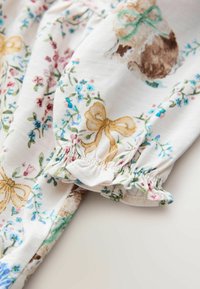 Auduma ar ziedu rakstu, kurā redzamas mazas zili un rozā ziedi, ar zelta loku akcentiem. Apkārt dūriens ar savilktu aproci, mīksts tekstūra un gaišas krāsas.