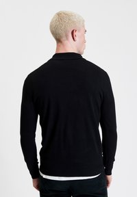 Zwarte langwerpige polo met lange mouwen en een kraag, gemaakt van een gemêleerde stof. Het heeft een aangesloten ontwerp en een rechtezoom. Draagt over een wit shirt.