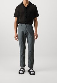 Maglietta nera a maniche corte con un design a maglia testurizzata e taschino sul petto; pantaloni grigi slim-fit; sandali neri con fibbie; calze bianche.