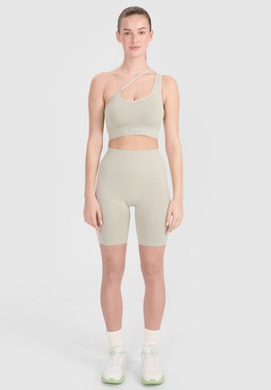 Kobieta stoi z neutralnym wyrazem twarzy, ubrana w beżowy crop top na jedno ramię oraz dopasowane wysokie spodenki rowerowe, białe sneakersy i skarpetki.