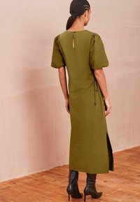 Rochie midi verde măslinie cu mâneci bufante, detalii pliate pe lateral și decupaj în formă de cheie la spate, asortată cu botine negre cu toc înalt până la genunchi.