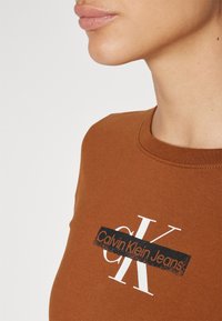 Hnědý bavlněný tričko s černobílým potiskem loga "Calvin Klein Jeans" přes hruď, s kulatým výstřihem.