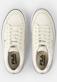 Sneakers in tela color crema con texture trapuntata, lacci bianchi e suola bordata di nero. Presentano il logo FILA marrone sui lati.