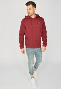 Maroonowy hoodie ze sznurkiem, boczną kieszenią na zamek i detalem logo, zestawiony z jasnymi zniszczonymi dżinsami i białymi trampkami.