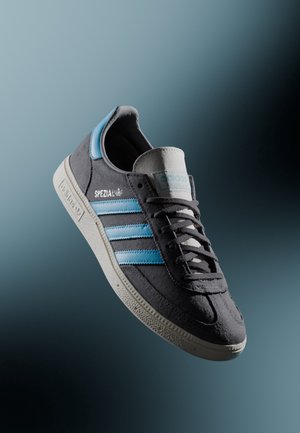Ciemnoszary sneaker Adidas Spezial z jasnoniebieskimi paskami i białą podeszwą na tle gradientu niebiesko-szarego.