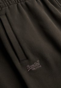 Pantalon de survêtement marron foncé avec taille élastique, poche latérale et logo SuperDry brodé avec des caractères japonais près de la poche.