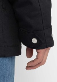 Morbida manica di giacca nera con polsino abbottonato, caratterizzata da una texture liscia e una leggera lucentezza. Mano poggiata accanto a un paio di jeans in denim.