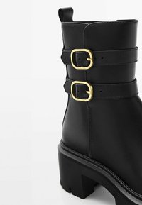 Stivaletto in pelle nera con due fibbie dorate, suola spessa e linguetta per facilitare l'indossamento. Texture liscia con dettagli di cucitura discreti.