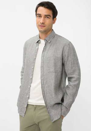 Mann trägt hellgraues Hemd über weißem T-Shirt mit olivgrüner Hose, steht mit einer Hand in der Tasche vor weißem Hintergrund.