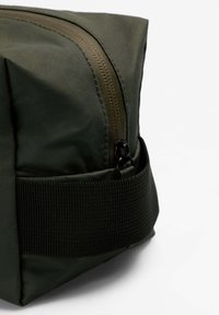 Bolsa de tela verde oscuro con cierre de cremallera, con una correa texturizada y forma cuadrada. Superficie lisa con detalles de costuras visibles.