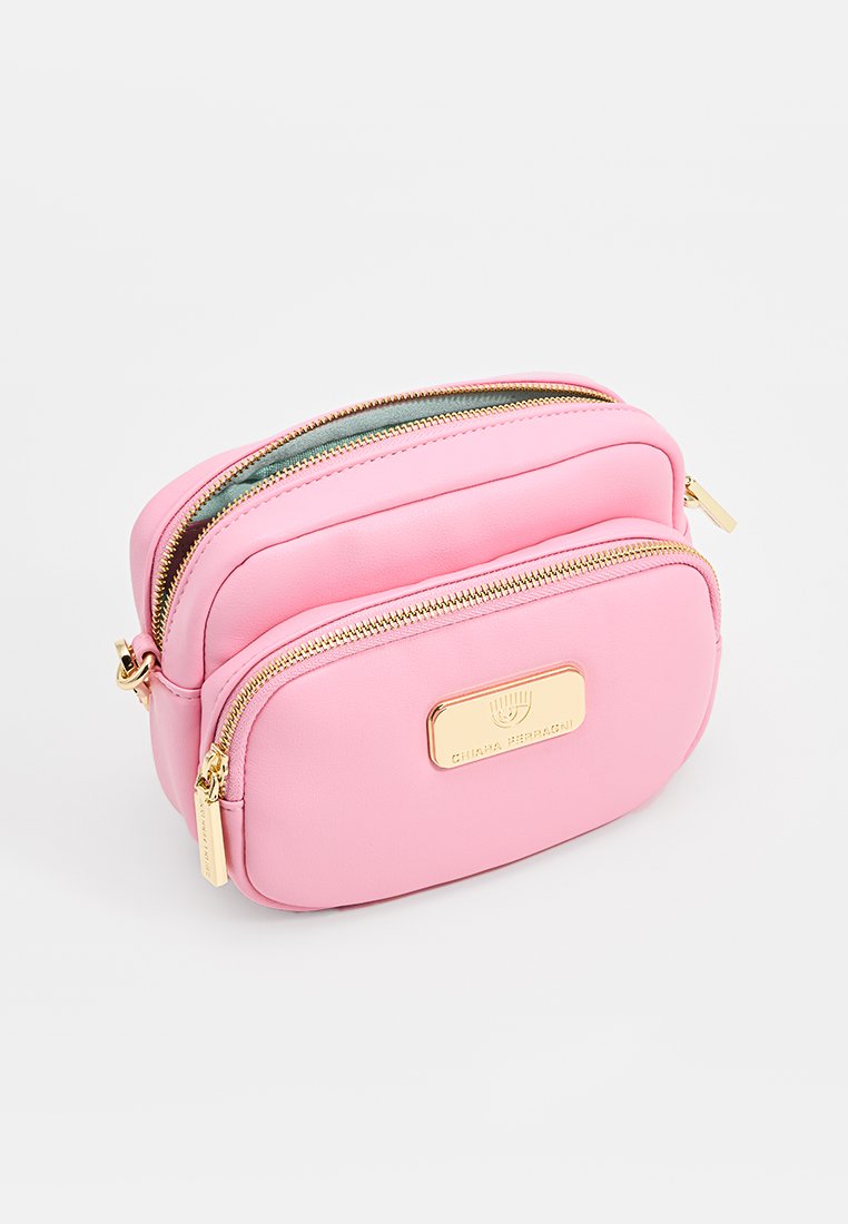 Różowa torebka na ramie w kształcie crossbody z dwoma złotymi zamkami błyskawicznymi i złotą plakietką z logo z przodu na jednolitym tle.