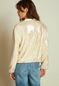 Sølvglitrende cremefarvet sweater med løs pasform og lange ærmer, der har en blød tekstur og iriserende pailletter, båret over blå denimjeans.