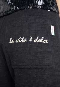 Pantalons en tissu texturé noir avec l'inscription brodée "la vita è dolce" en crème au dos, avec une petite étiquette de marque dans le coin.