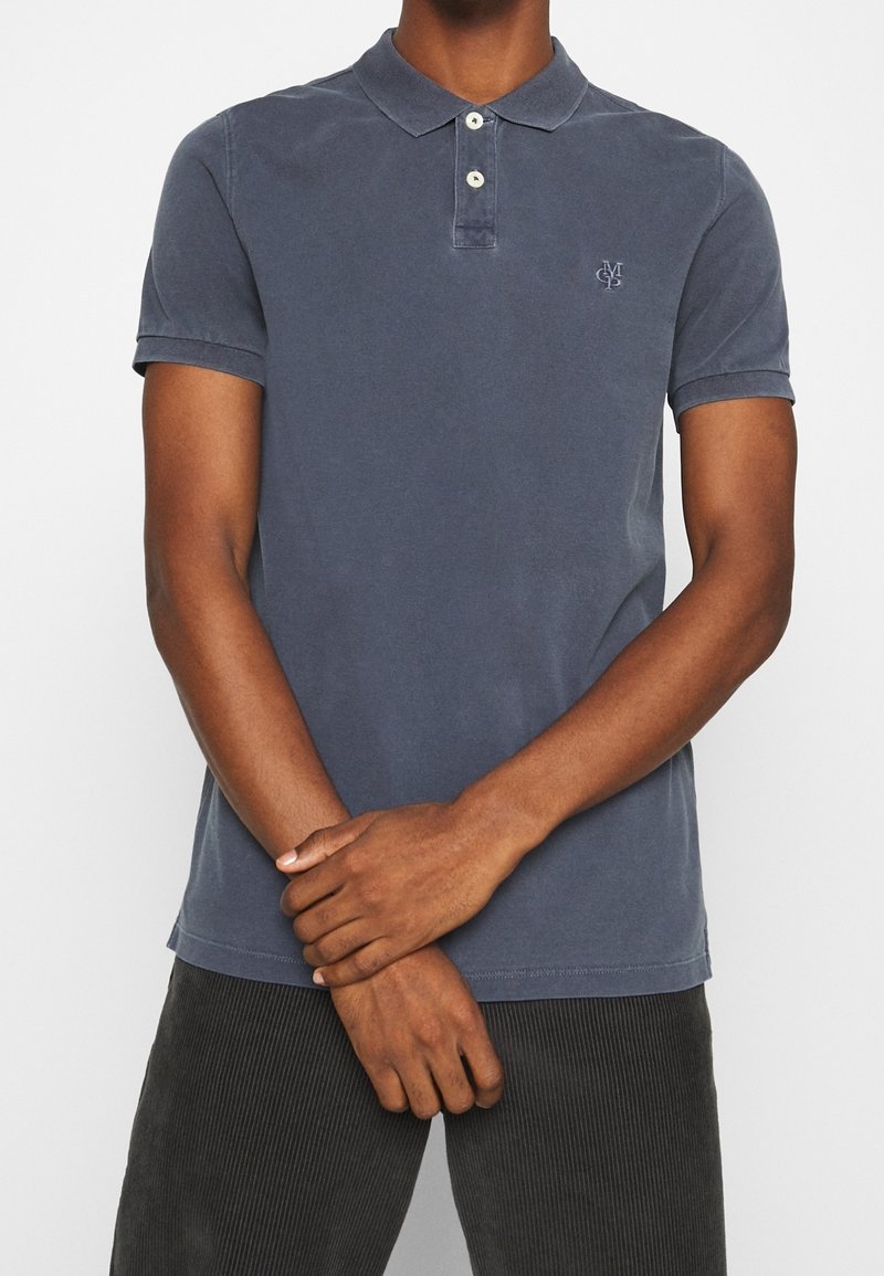 Homme portant un polo bleu délavé à manches courtes avec deux boutons et un petit logo brodé sur la poitrine, debout, les mains croisées.