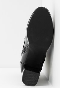 Chaussure en cuir noir avec une semelle texturée, dotée d'un bout arrondi et d'un petit talon. Présentée en angle sur un fond blanc.