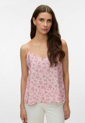 Ung kvinde med langt brunt hår iført en lyserød floral spaghetti-strop top og hvide bukser, stående mod en enkel baggrund.