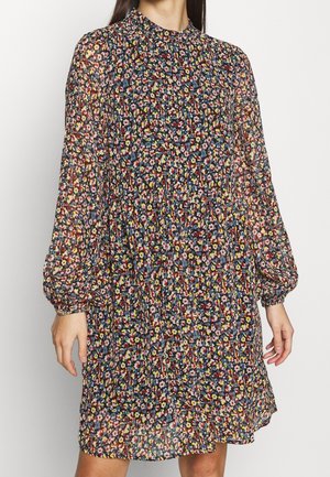 Mujer con un vestido de manga larga y cuello alto con un pequeño patrón floral de colores en rojo, amarillo, azul y rosa sobre un fondo oscuro.