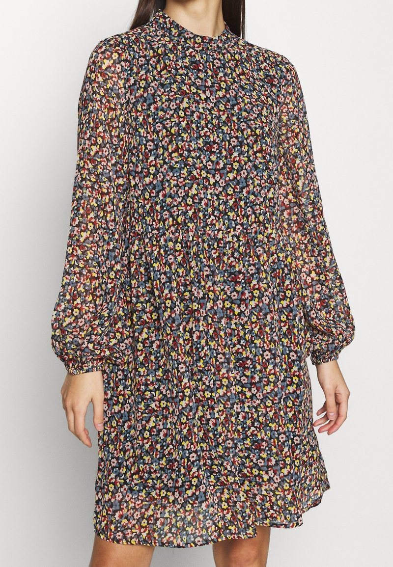 Femme portant une robe à manches longues et col montant avec un petit motif floral coloré en rouge, jaune, bleu et rose sur un fond sombre.