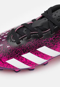 Primer plano de unas botas de fútbol negras y rosas con superficie texturizada, cordones negros y tres franjas blancas en el costado.