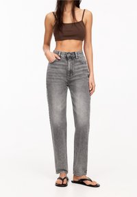 Femme portant un jean gris taille haute, un débardeur court marron et des sandales plates noires, debout avec une main dans la poche.