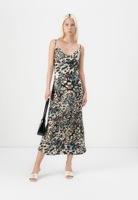 Woman with long blonde hair wearing a sleeveless animal print midi dress, white open-toe heels, and holding a black handbag.
