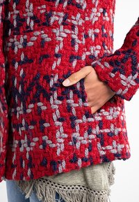 Mano in tasca di una giacca rossa a trama testurizzata con motivo intrecciato blu e grigio, sopra un top grigio con frange e jeans blu.