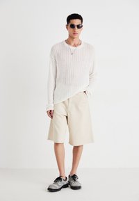 Lekki sweter z prążkowanej kremowej dzianiny, beżowe dopasowane szorty i czarne buty na sznurówki z teksturalnym detalem; wygląd dopełniają okrągłe okulary przeciwsłoneczne.