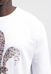 Bílé tričko s dlouhým rukávem s barevným, texturovaným abstraktním motivem fleur-de-lis na přední straně, s hladkým bavlněným povrchem.