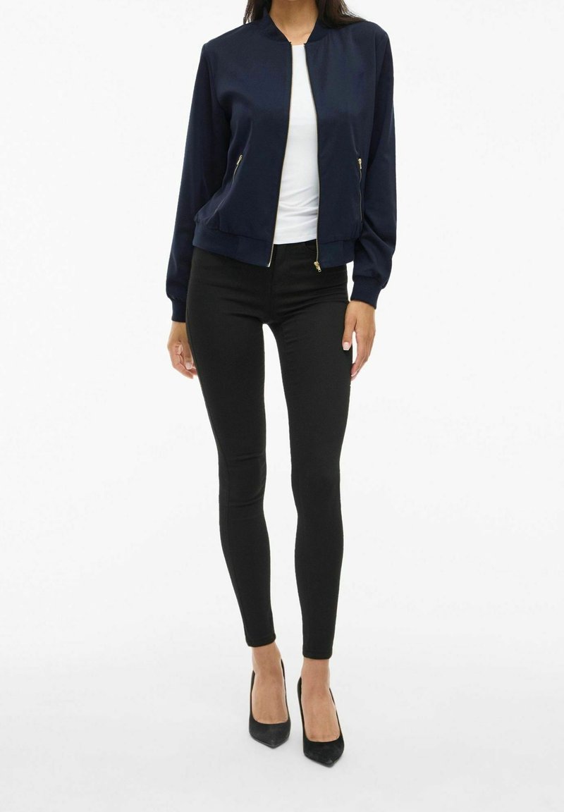 Femme portant une veste zippée bleu marine, une chemise blanche, un pantalon noir slim et des escarpins noirs pointus, debout devant un fond blanc.