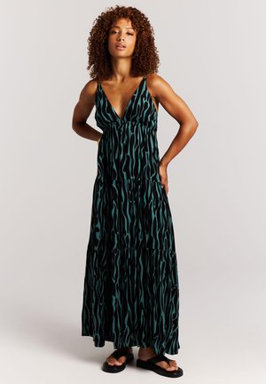 Vrouw met krullend haar, gekleed in een mouwloze maxi-jurk met een groenblauw en zwart patroon en zwarte sandalen, staand met handen in haar zij.