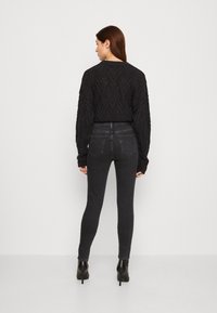 Μαύρο πλεκτό πουλόβερ με υφασμάτινο σχέδιο ζιγκ-ζαγκ, συνδυασμένο με σκούρο τζιν. Το μοντέλο φορά μαύρες γόβες με τακούνι πάνω από τον ασ�τράγαλο. Η εικόνα δείχνει την πίσω όψη.