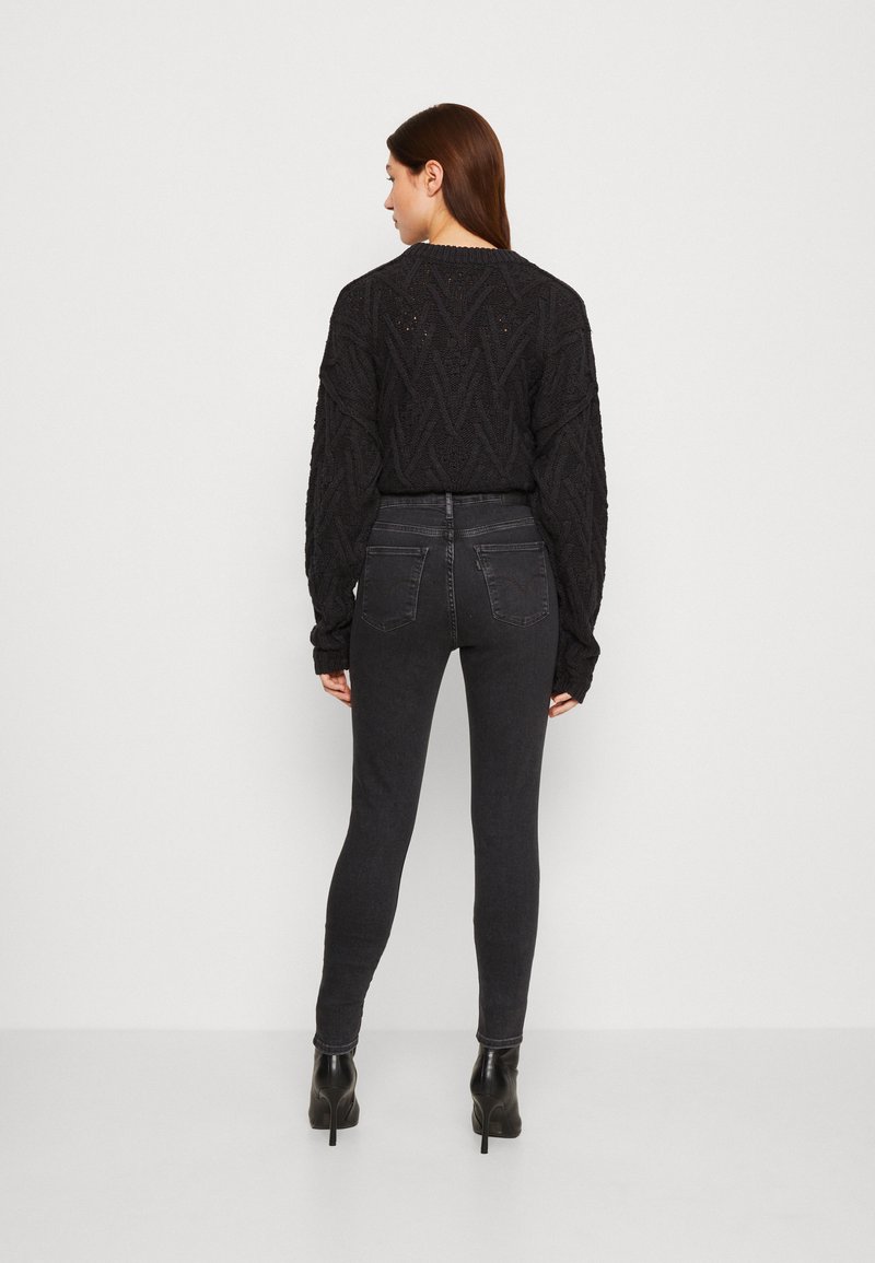 Pull en maille noire avec un motif zigzag texturé, associé à un jean foncé. Le mannequin porte des bottines à talons noires. L'image montre la vue arrière.