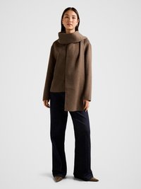 Ruda vilnonė palaidinė su plačiu, drapiruotu apykakliu, derinama su tamsiai mėlynomis plačiomis kelnėmis ir smėlio spalvos smailiais bateliais. Paprastos, švarios linijos.