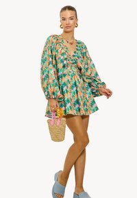 Vestido de estampado floral verde con escote rizado y mangas largas abullonadas. Acentuado con un bolso de mimbre y unos zapatos azulones sin cierres.