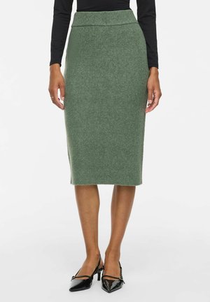 Mujer con falda lápiz de punto verde hasta la rodilla combinada con top negro de manga larga y tacones negros de punta con tiras.