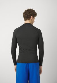 Langermet svart treningstopp med en tettsittende design, glatt tekstur og raglanermer. Komplimentert av blå shorts.