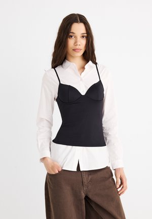 Młoda kobieta o długich włosach, ubrana w białą koszulę na guziki, na którą nałożona jest dopasowana czarna koszulka na ramiączkach i luźne brązowe spodnie, pozująca na białym tle.