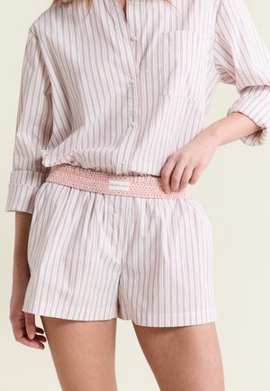 Person trägt weiß-rosa gestreiftes Langarmshirt und passende Shorts, mit einem rosa elastischen Gürtel mit der Aufschrift "Healthy soul" um die Taille.