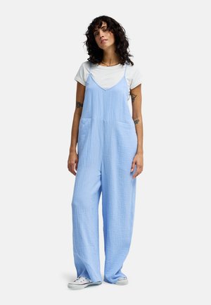Lichtblauw katoenen jumpsuit met een gestructureerde afwerking, voorzien van verstelbare bandjes en twee zakken aan de voorkant, gecombineerd met een wit T-shirt en sneakers.