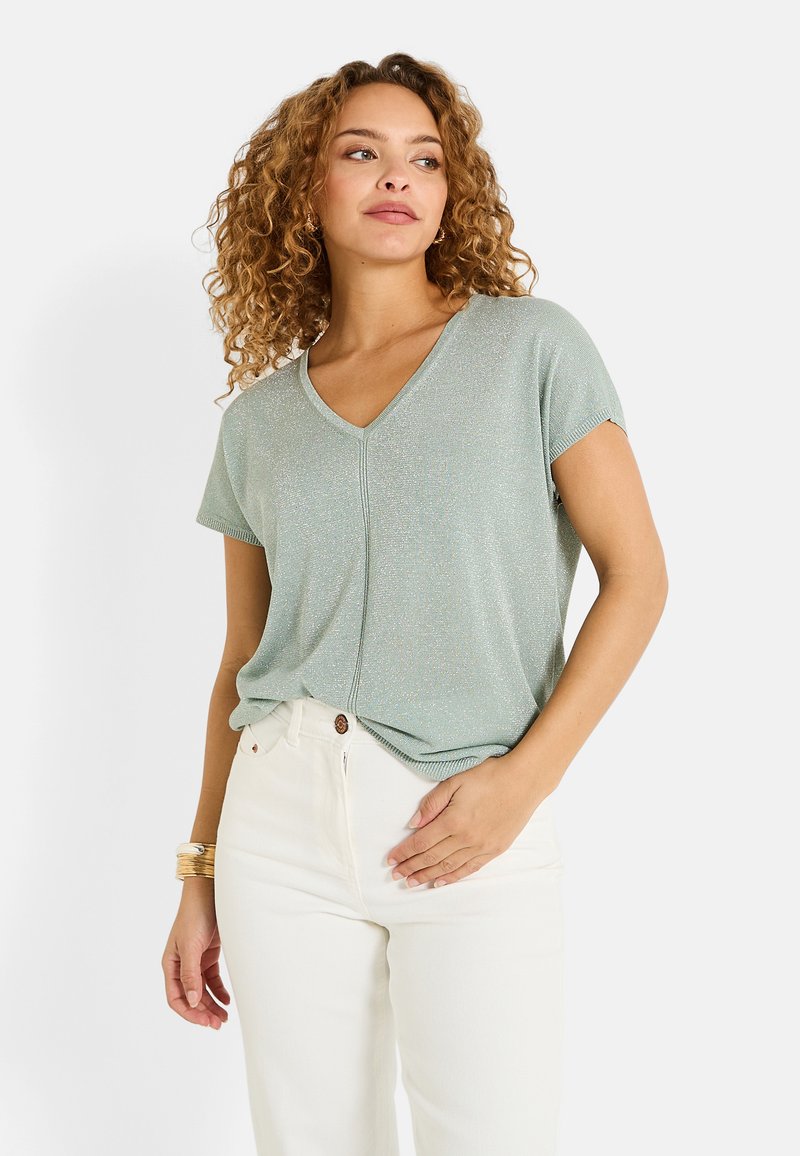 Top de manga corta en verde menta claro con un sutil brillo, que presenta un corte relajado y un detalle de costura central. Llevado con jeans blancos.