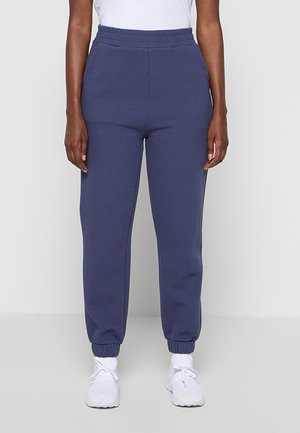 Personne portant un pantalon de jogging en maille texturée bleu marine avec taille et poignets élastiques, assorti à des baskets blanches et un haut blanc.
