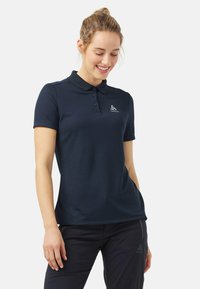 Navyblauwe polo met een kraag, drie knopen en een logo. Gemaakt van gladde stof met een getailleerde snit, gecombineerd met donkere broeken.