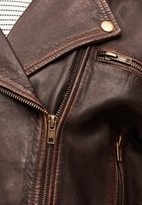 Κοντινό πλάνο καφέ δερμάτινου μπουφάν που δείχνει το μεταλλικό φερμουάρ, την τσέπη με φερμουάρ και το ραμμένο γιακά με χάλκινο κουμπί snap.