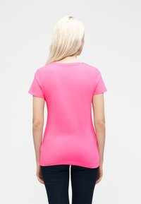 Γυναίκα με μακριά ξανθά μαλλιά, φορώντας ένα φωτεινό ροζ κοντομάνικο μπλουζάκι και σκούρο παντελόνι, στέκεται με την πλάτη γυρισμένη σε λευκό φόντο.