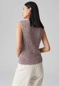 Maglione senza maniche aderente in maglia testurizzata color bordeaux e crema. Caratterizzato da un collo alto e bordo a costine. Indossato con pantaloni chiari.