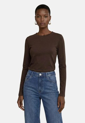 Kvinde iført en tætsiddende mørkebrun langærmet top med subtil bølget halsudskæring og blå højtaljede jeans, stående mod en ensfarvet baggrund.